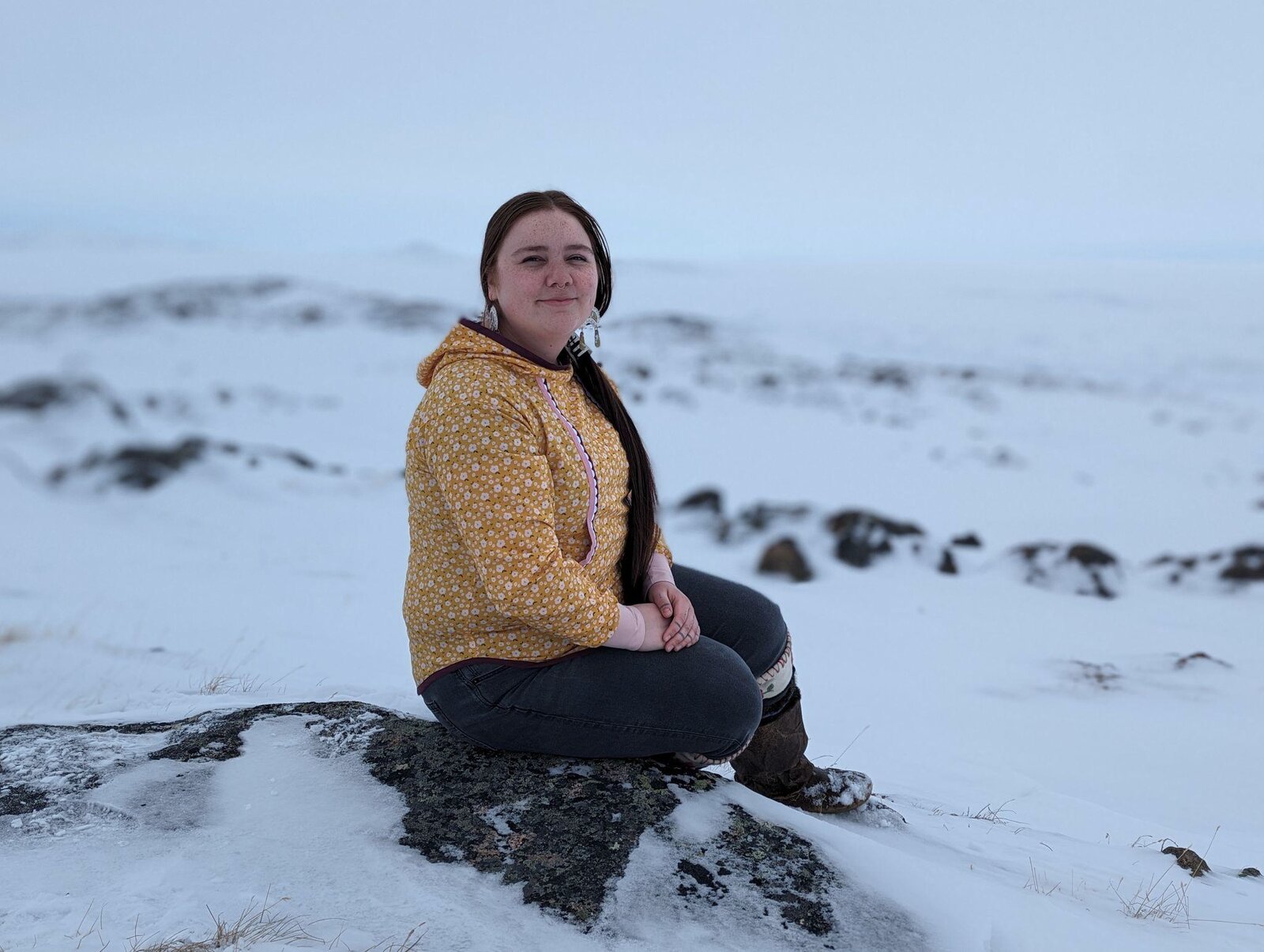 Erica sitting on a rock in a snowy coastal landscape, wearing a yellow sweater