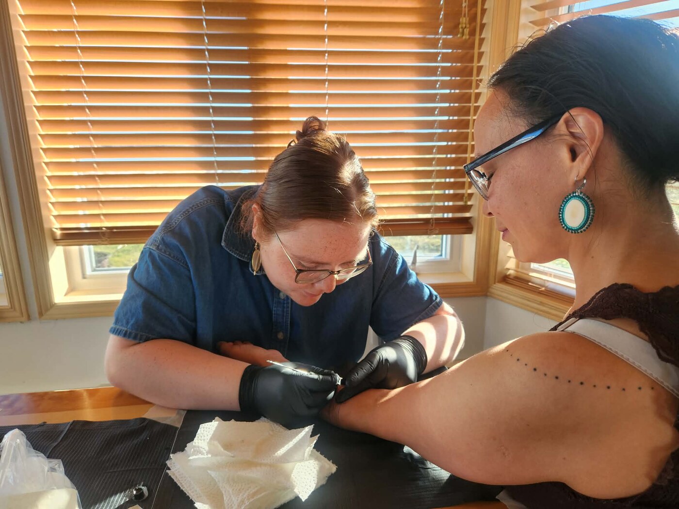 Erica, wearing gloves and glasses, leaning in to hand-poke a client's arm with a stick-and-poke tattoo needle, sunlight through the window behind her
